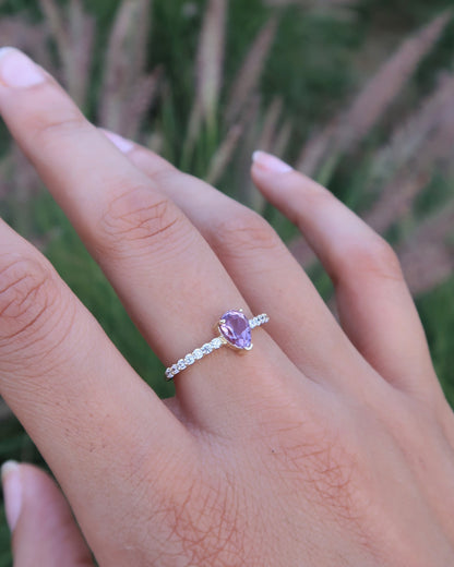 Hand wearing a ring with a purple gemstone against a blurred green background