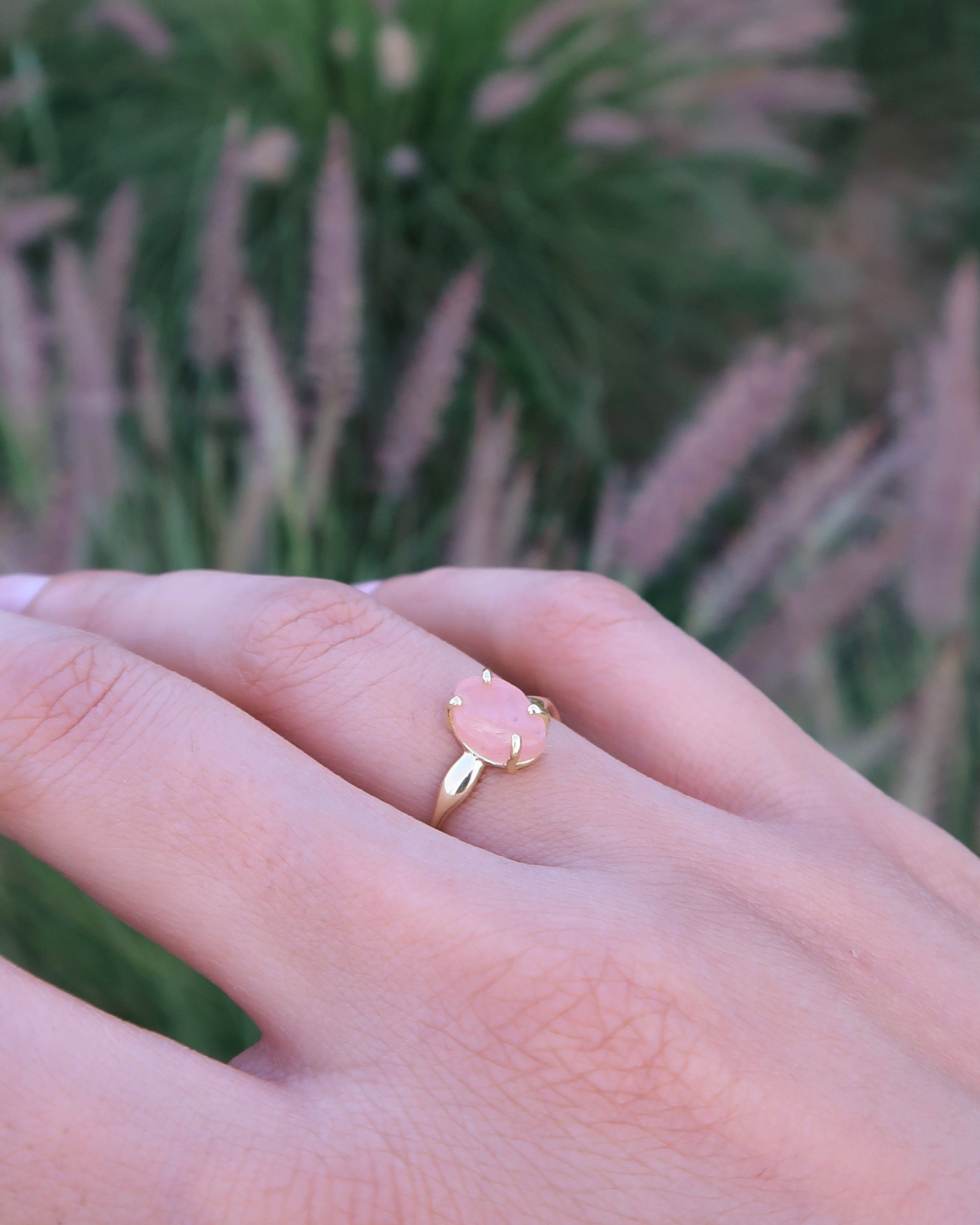 Hand wearing a gold ring with a pink gemstone in front of a blurred background of tall grasses.