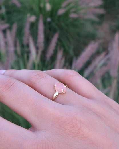 Hand wearing a gold ring with a pink gemstone in front of a blurred background of tall grasses.