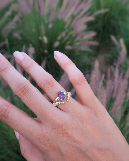 Hand wearing a ring with a purple gemstone against a blurred green background