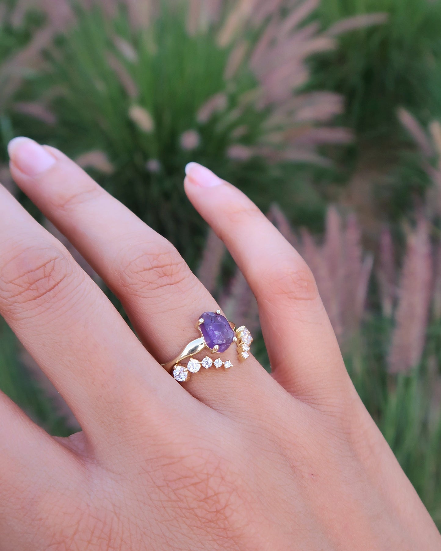 Hand wearing a ring with a purple gemstone against a blurred natural background
